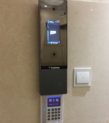 A wall-mounted face recognition device with a screen above a numeric keypad and a nearby light switch on a beige wall.